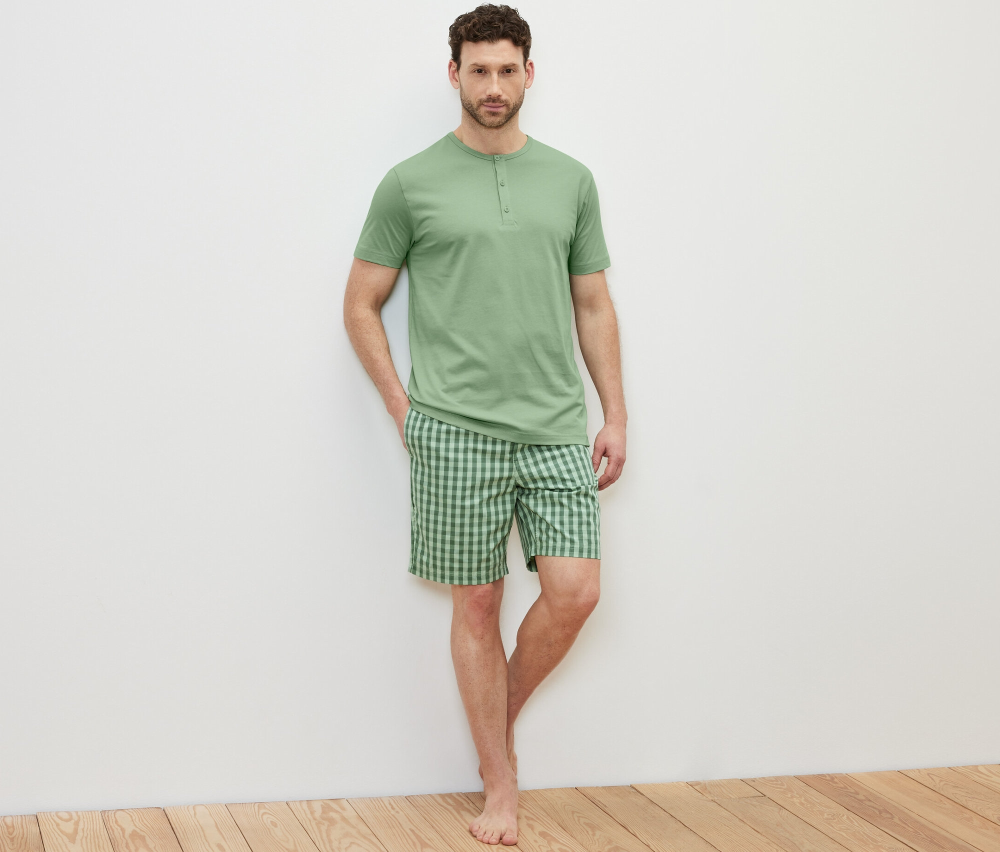 Açık renkli bir arka planda şortlu pijama takımı giyen bir adam poz veriyor.