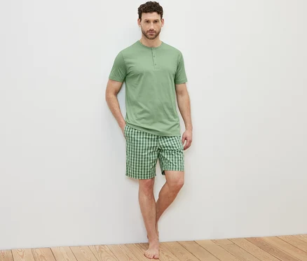 Açık renkli bir arka planda şortlu pijama takımı giyen bir adam poz veriyor.