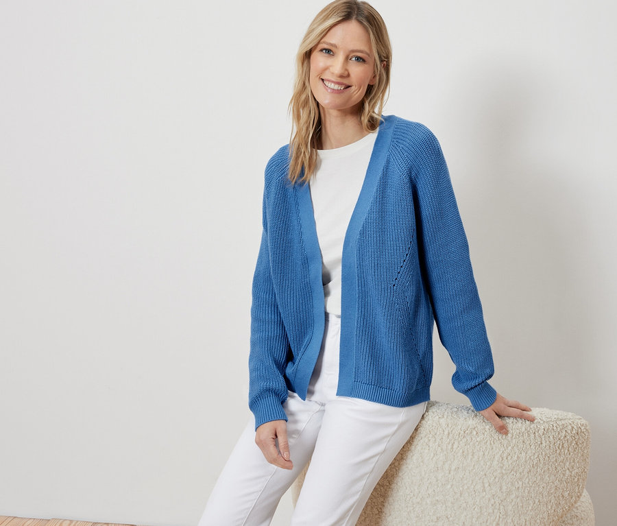 Kadın oturuyor ve mavi, açık bir hırka giyiyor.