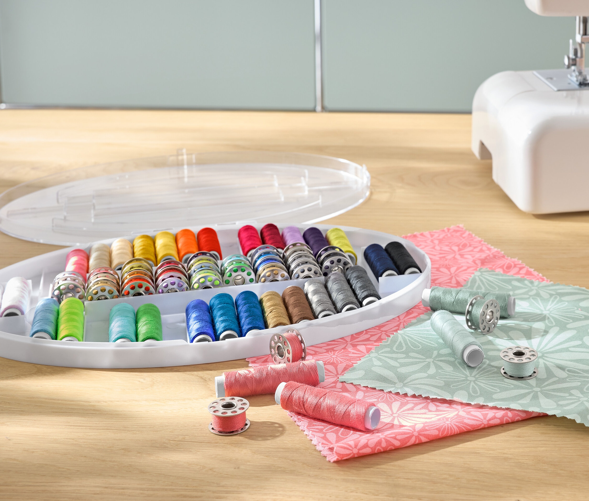 Plastik bir düzenleyicideki renkli iplik makaraları ile bir dikiş ipliği seti, bir dikiş makinesi ve kumaşların yanında.