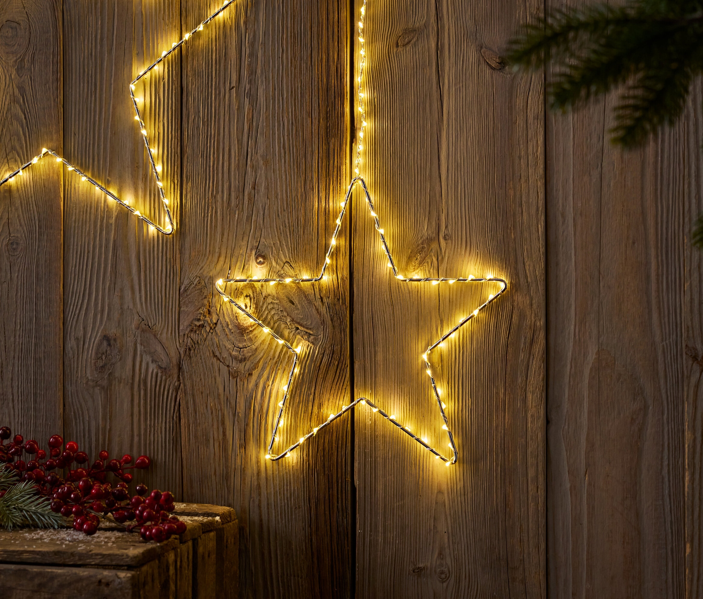 Ahşap bir arka plan üzerinde, çam dalı ve kırmızı meyvelerle birlikte iki adet ışıklı yıldız.