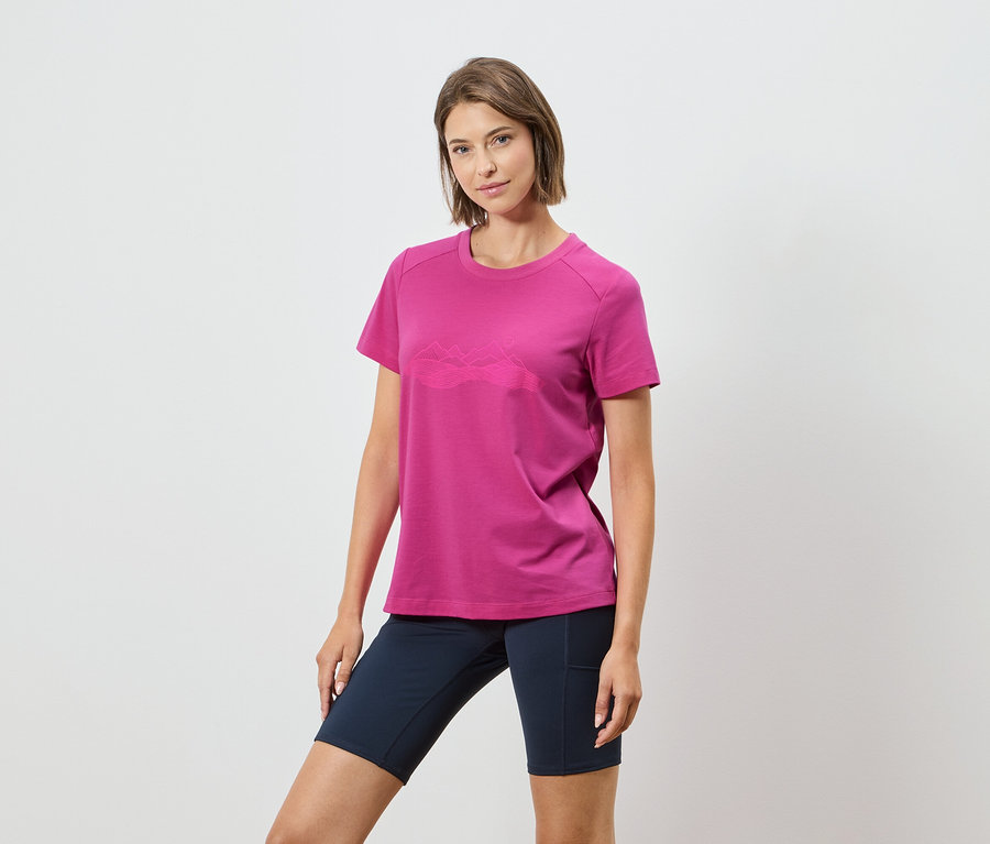 Kadın beyaz bir arka planda dağ sırası olan pembe bir tişört ve koyu mavi şort giyiyor.