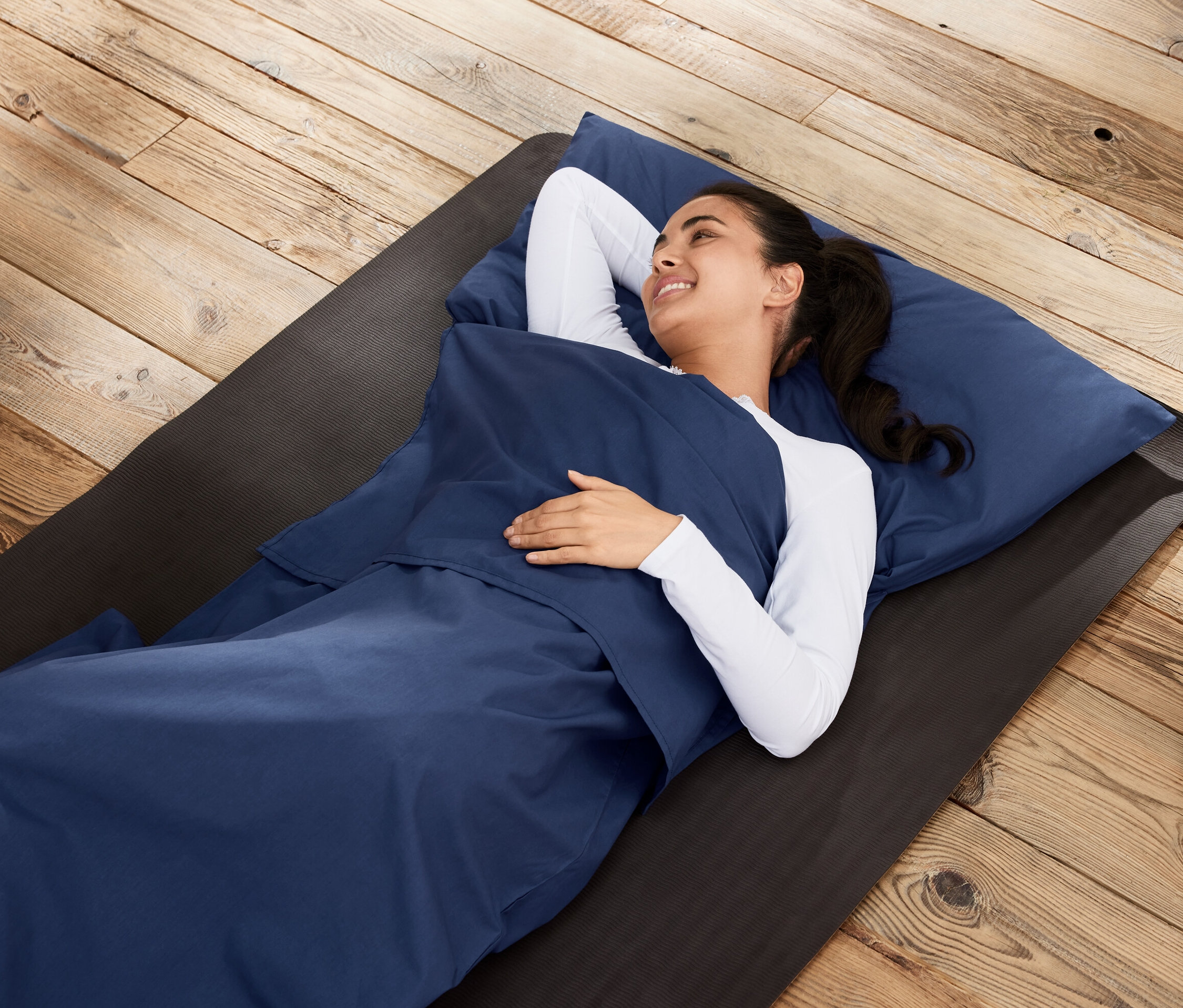 Kadın, ahşap zemindeki bir matın üzerinde uyku tulumu içliği içinde yatıyor.