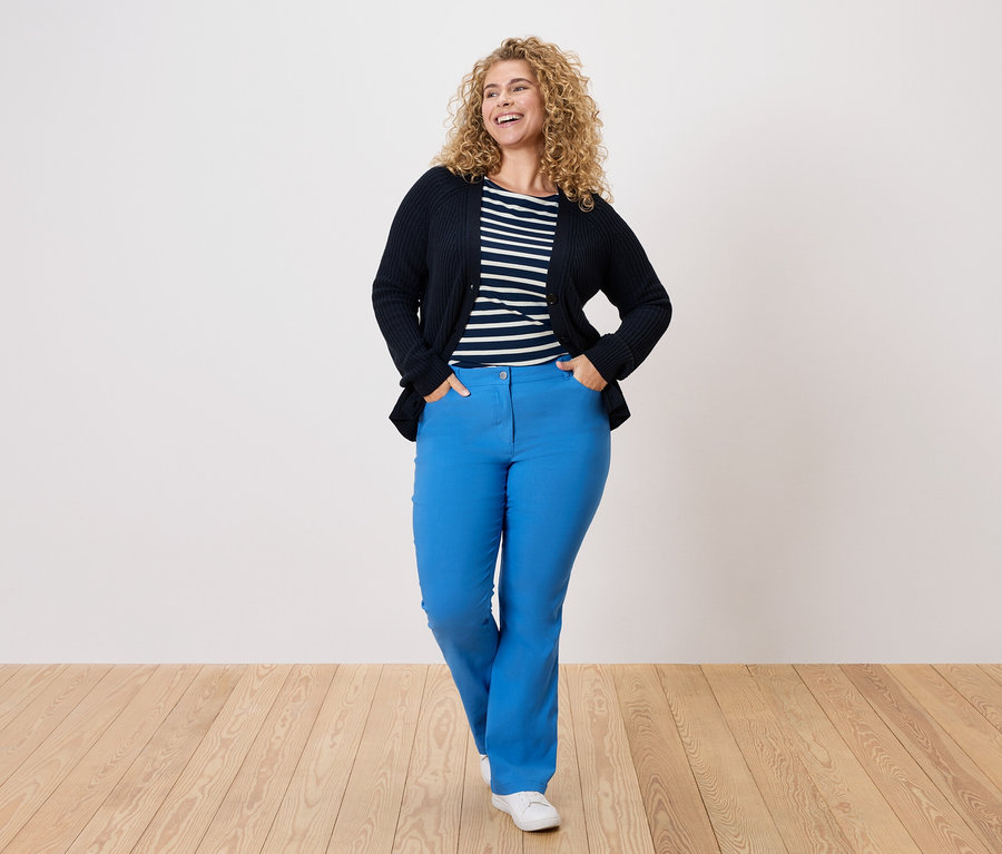 Kıvırcık saçlı kadın lacivert bir hırka, çizgili bir gömlek ve mavi pantolon giyiyor.