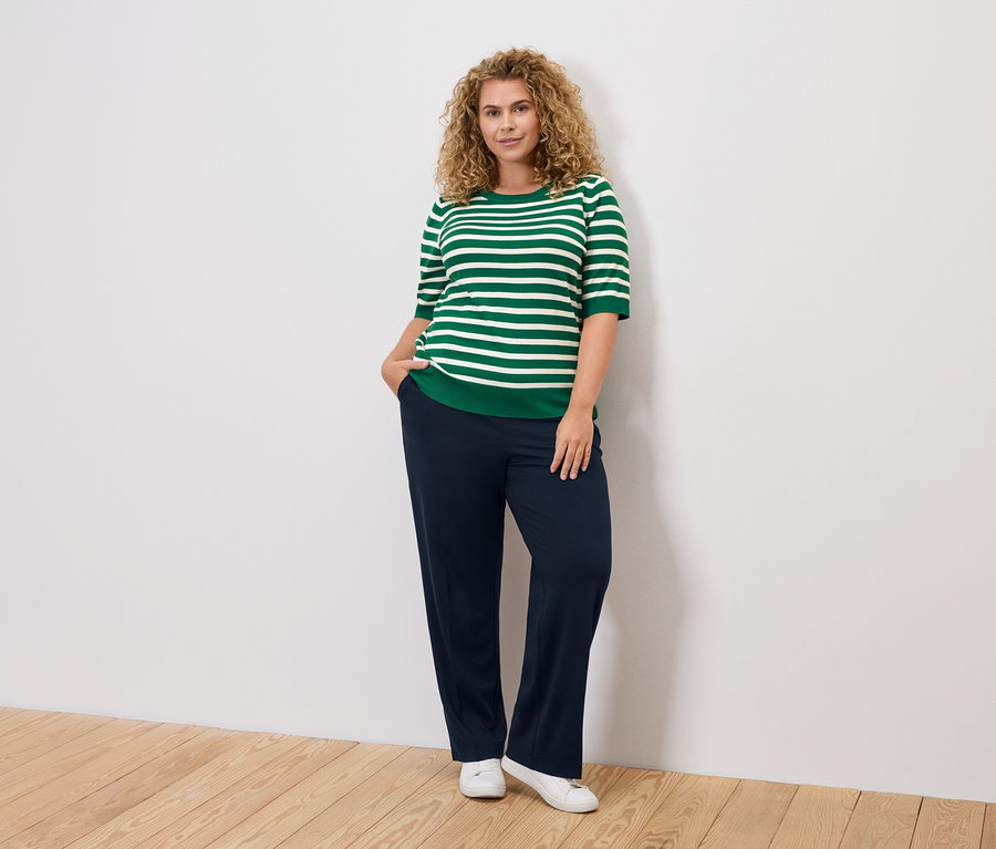 Kıvırcık saçlı bir kadın, yeşil beyaz çizgili bir üst, lacivert pantolon ve beyaz ayakkabılar giyiyor.