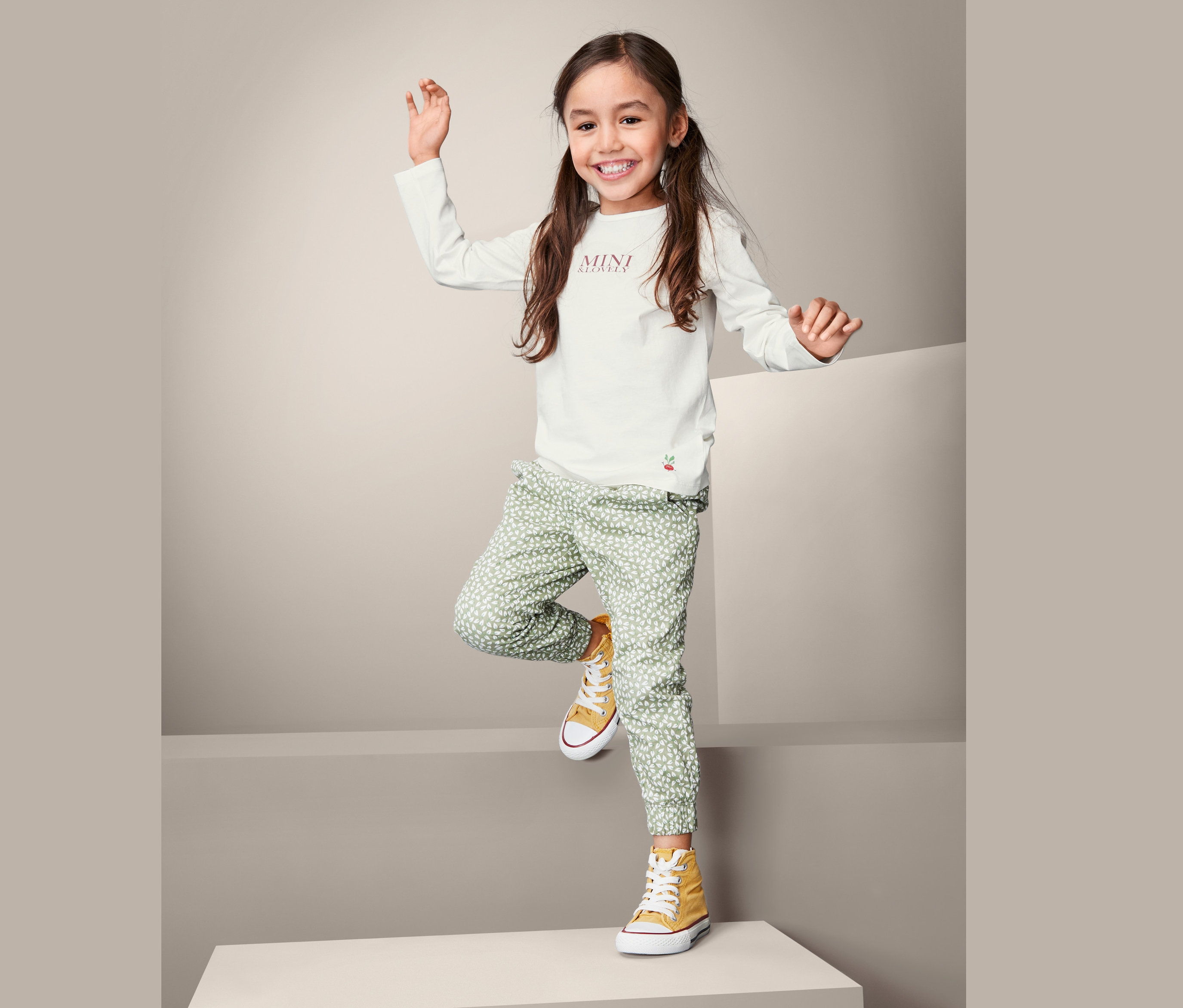 Gülümseyen kız uzun kollu tişört, yeşil desenli pantolon ve sarı spor ayakkabılarla poz veriyor.