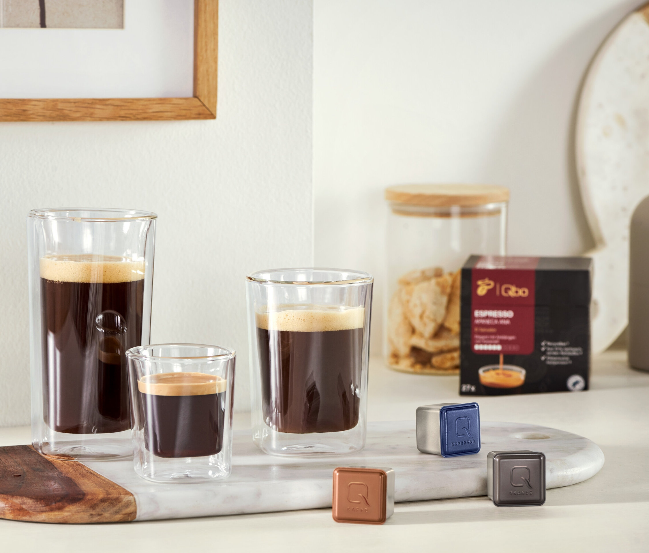 Mermer bir tepsi üzerinde, yanında bir kapsül paketi ve üç kahve kapsülü bulunan, farklı boyutlarda üç kahve bardağı duruyor. 2 Caffè Crema Bardağı ve 2 Espresso Bardağı görülüyor.