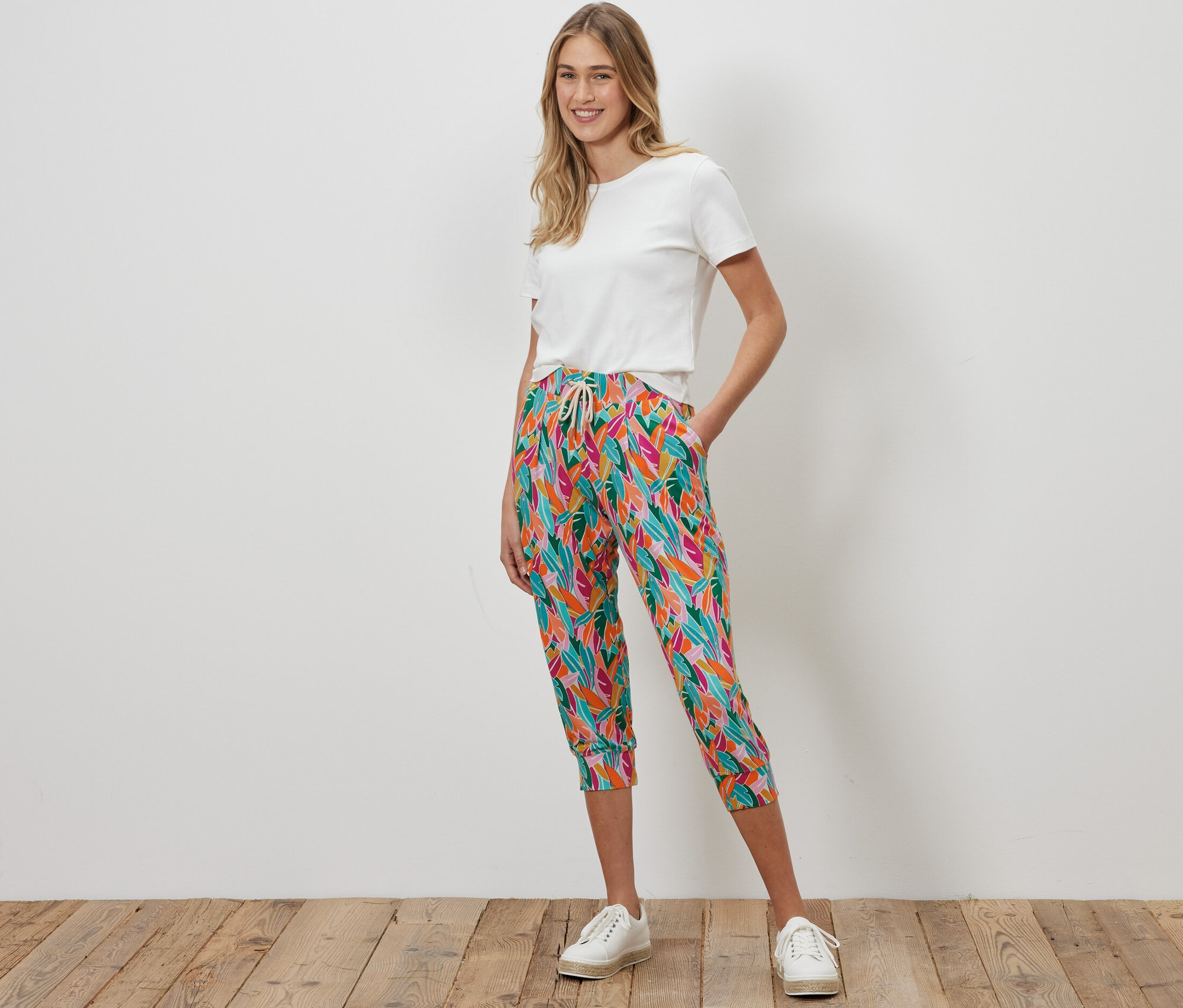 Bir kadın ayakta duruyor ve çok renkli 3/4 jarse pantolon ve beyaz bir tişört giyiyor.