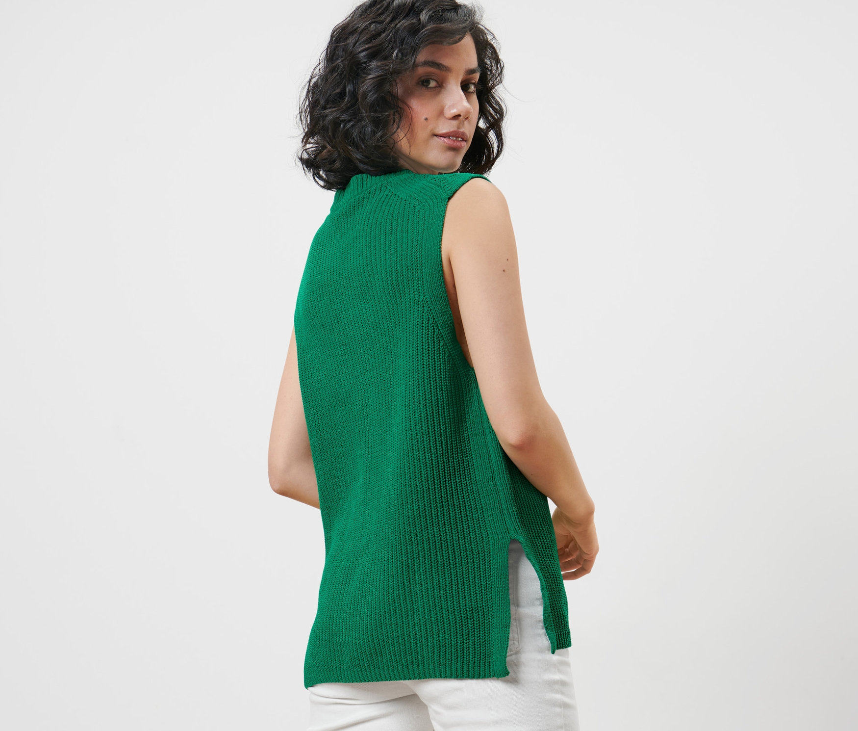 Omuzunun üzerinden bakan kadın, yeşil kalın örgü kolsuz süveter ve beyaz pantolon giyiyor.