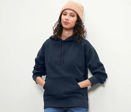 Kahverengi kıvırcık saçlı ve bereli bir kadın, lacivert kapüşonlu bir sweatshirt ile poz veriyor.