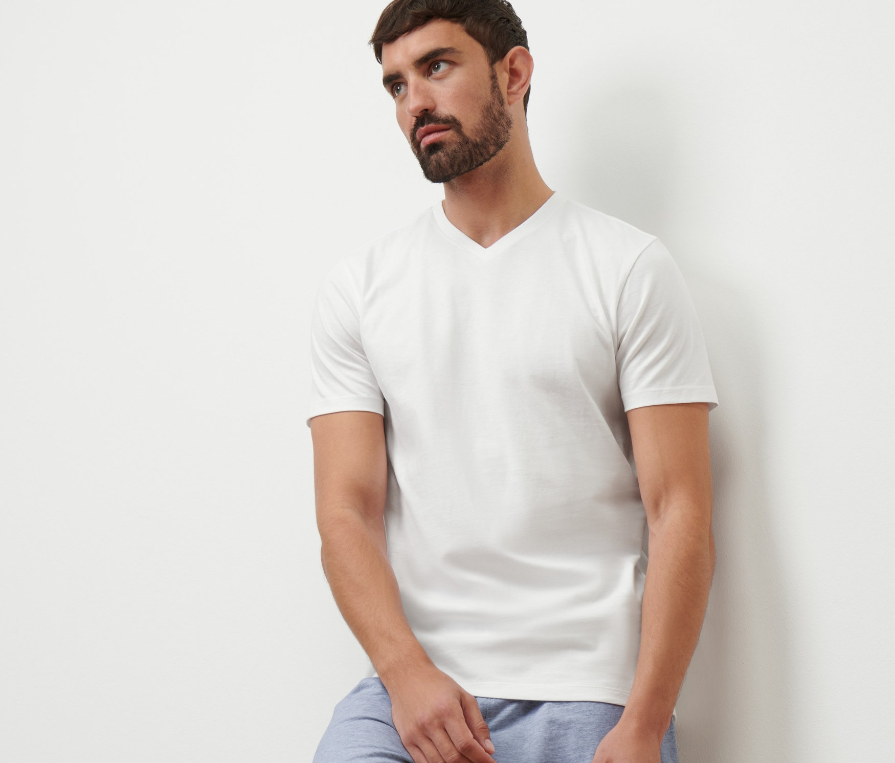 Duvara yaslanmış, beyaz V yaka tişört ve mavi şort giyen bir adam.