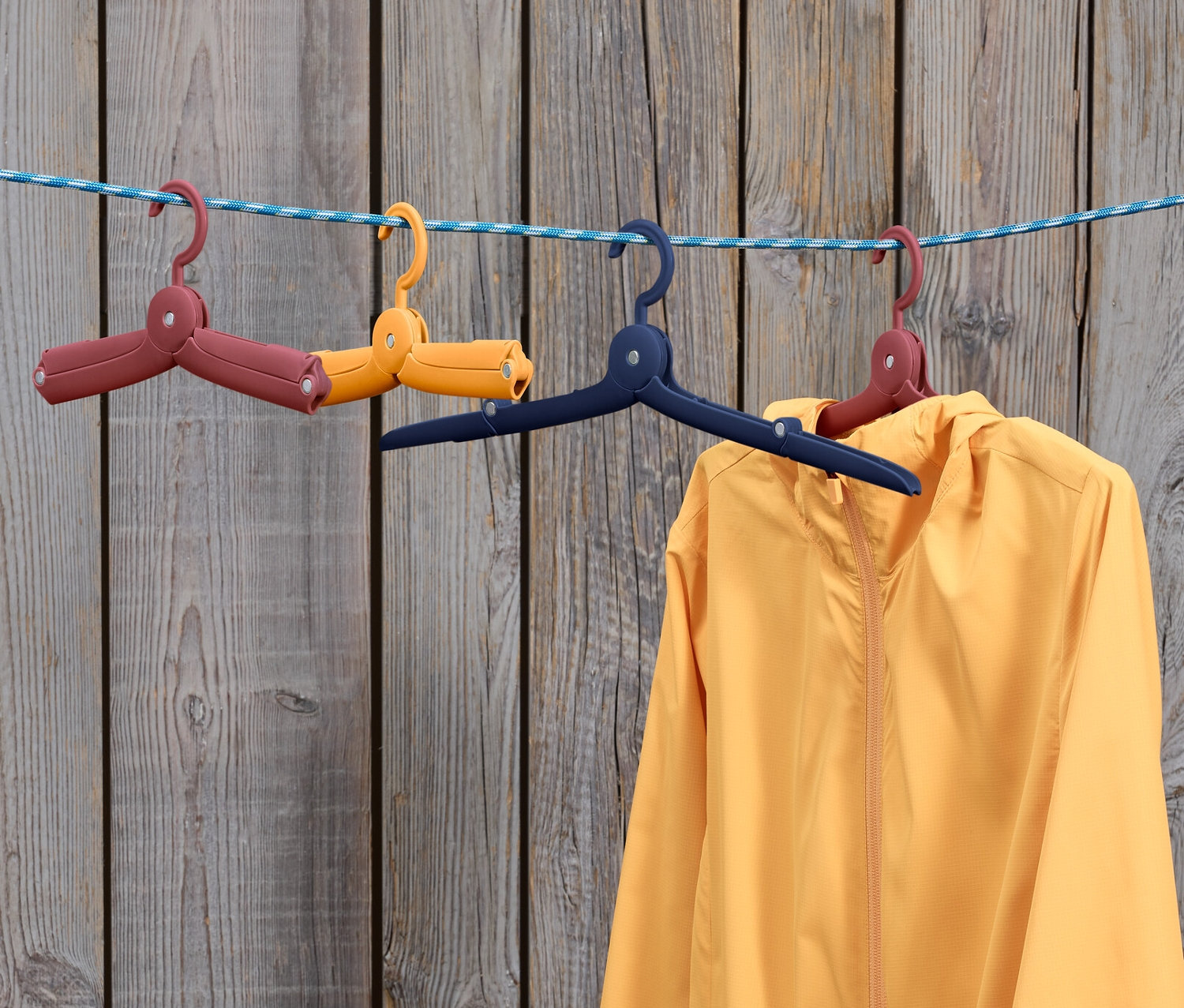 İpte mavi askıda sarı bir ceket asılı duruyor, yanında katlanmış iki adet 6 Adet Katlanabilir Yolculuk Askısı: kahverengi ve sarı.