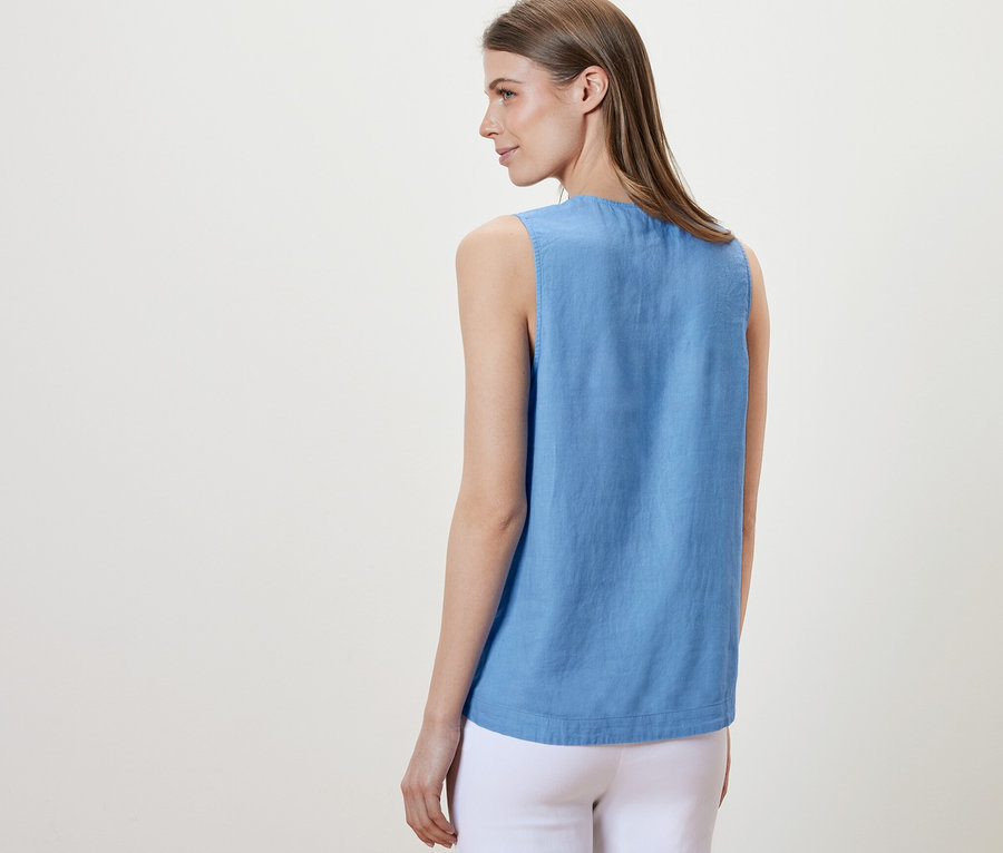 Kadın kameraya arkası dönük duruyor ve açık mavi, kolsuz bir keten bluz giyiyor. 