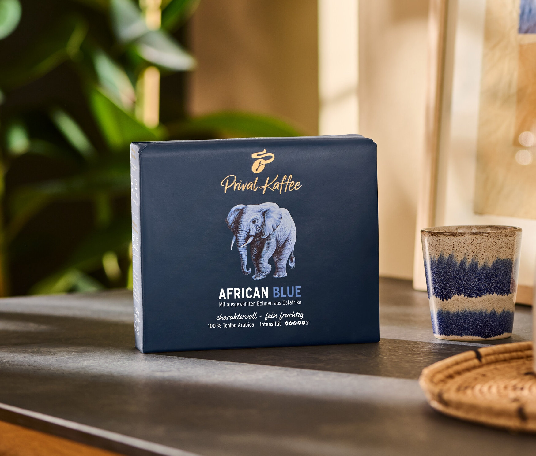 Bir paket Privat Kaffee African Blue - 500 g Öğütülmüş, bir fincan ve bir sürahi yanında bir masanın üzerinde duruyor.