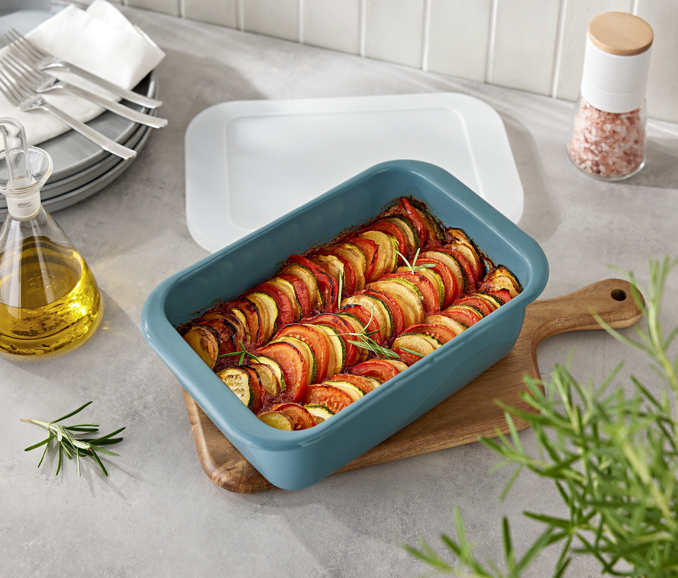 Ahşap bir tahtada mavi bir fırın kabında sebzeler. Yanında bir şişe yağ ve baharatlık.