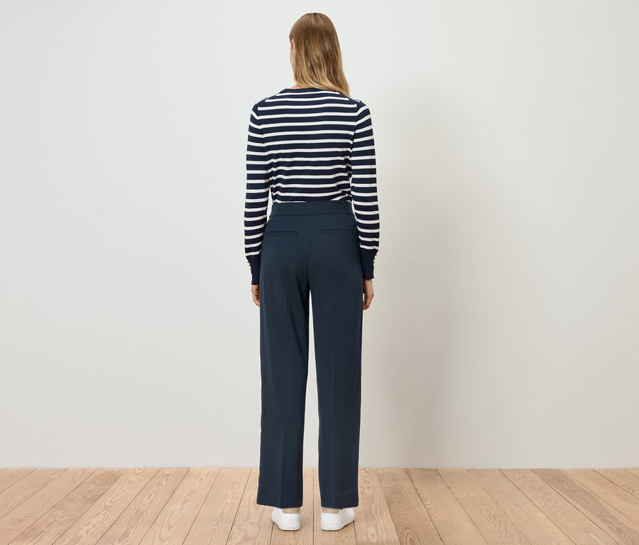 Bir kadın arkası dönük duruyor, üzerinde mavi pantolon ve çizgili bir üst var.