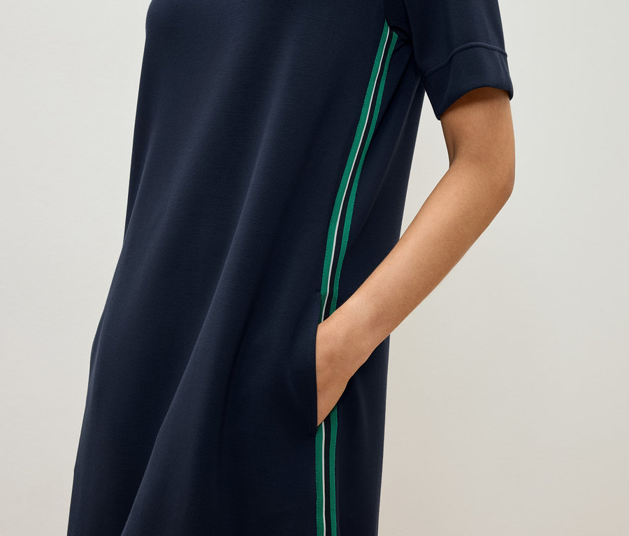 Kadın, yeşil ve beyaz şeritli, lacivert, kısa kollu bir elbisenin cebinde elini tutuyor.