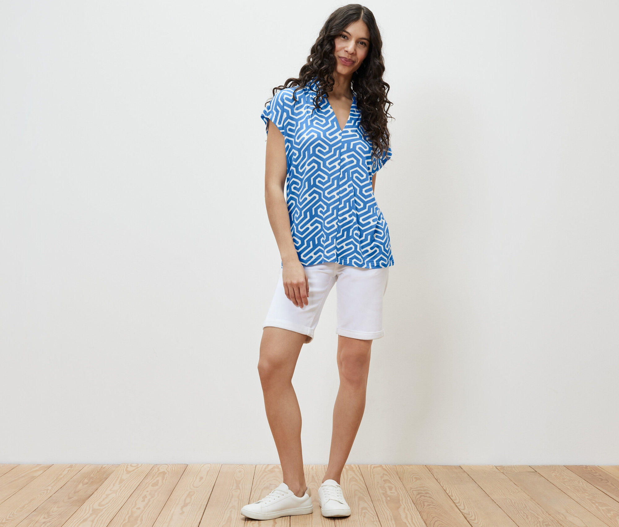 Bir kadın ayakta duruyor, mavi V yakalı bir bluz ve beyaz şort giyiyor.