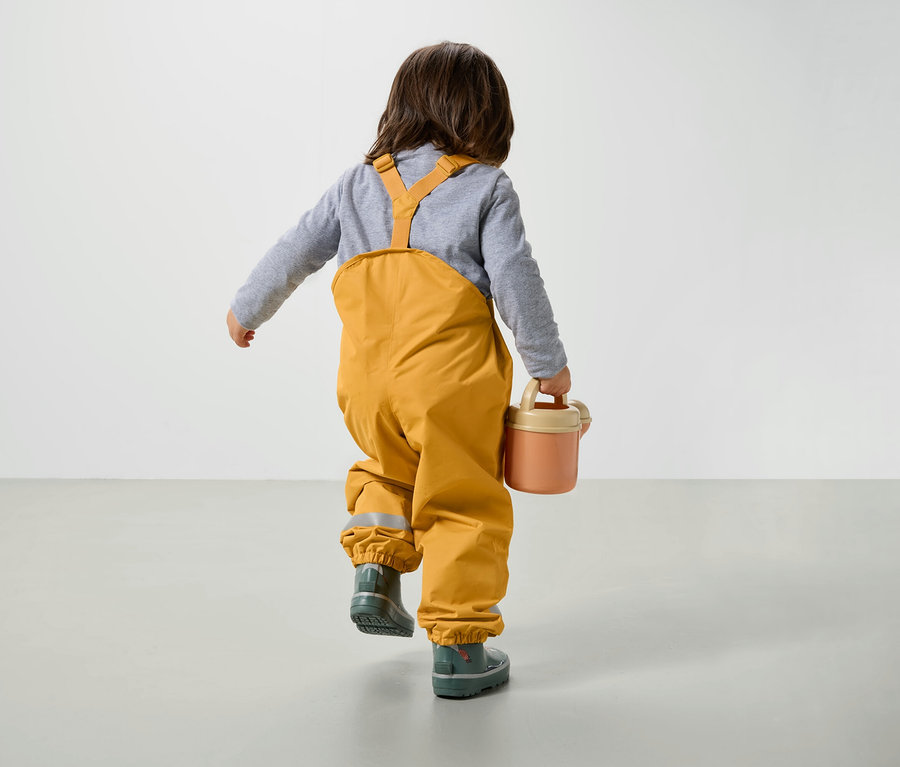 Sarı tulum ve lastik çizme giyen bir erkek çocuk yerde yürüyor ve pembe bir sulama kabı taşıyor.