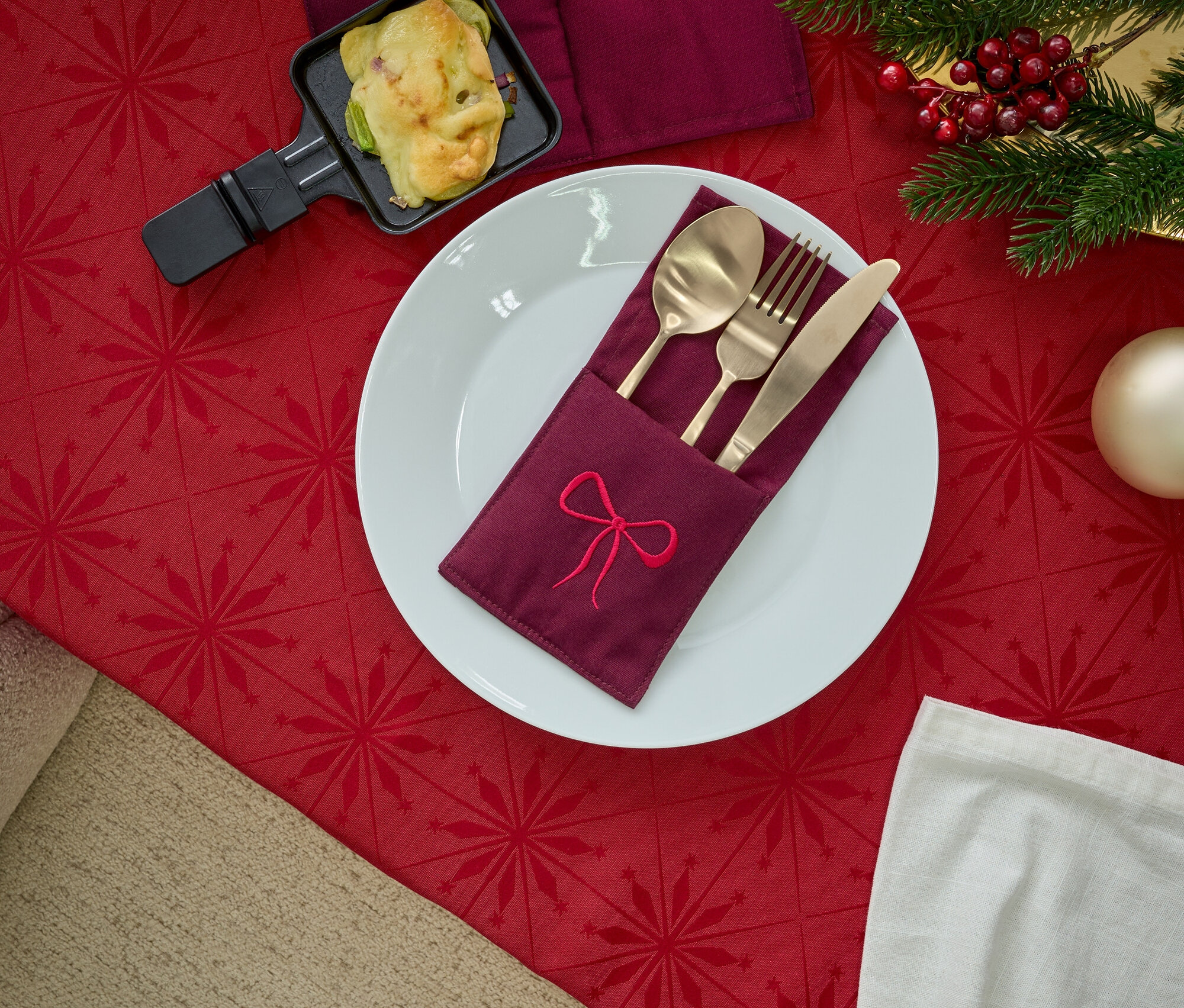 Kırmızı masa örtüsü üzerinde işlemeli peçetelik, altın çatal bıçak takımı, raclette tavası ve Noel dekorasyonları bulunan tabak.