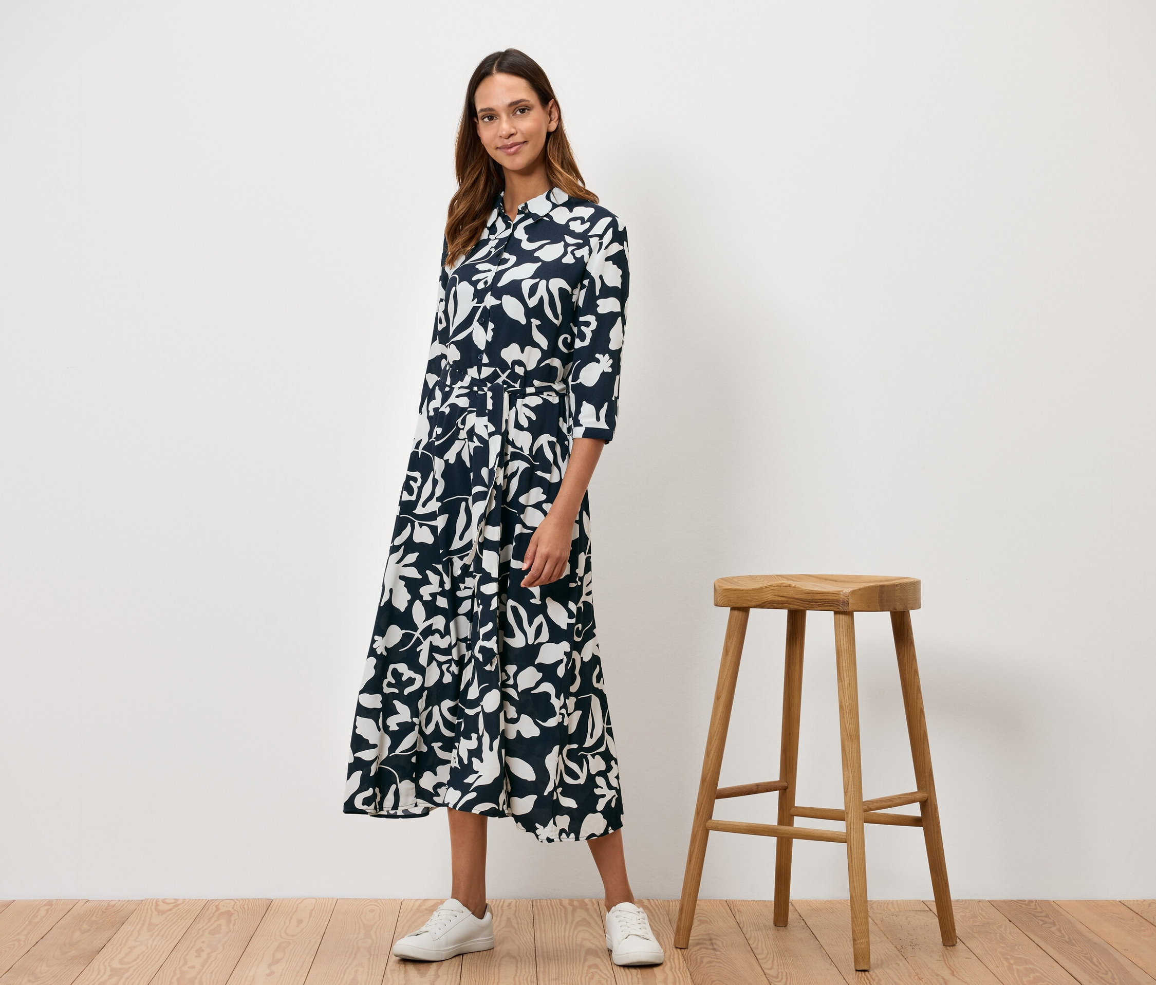 Bir kadın ahşap bir taburenin yanında duruyor ve beyaz çiçek desenli lacivert bir elbise giyiyor.