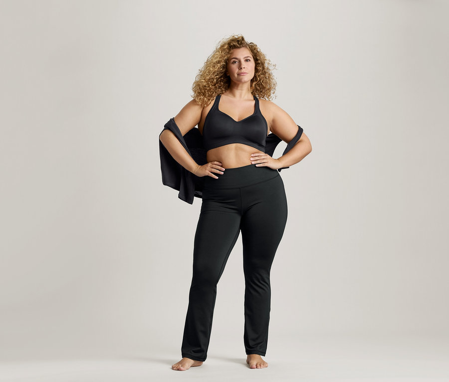 Kıvırcık saçlı, siyah sporcu sütyeni ve tayt giyen, omuzlarında ceket olan ve elleri kalçasında duran kadın.