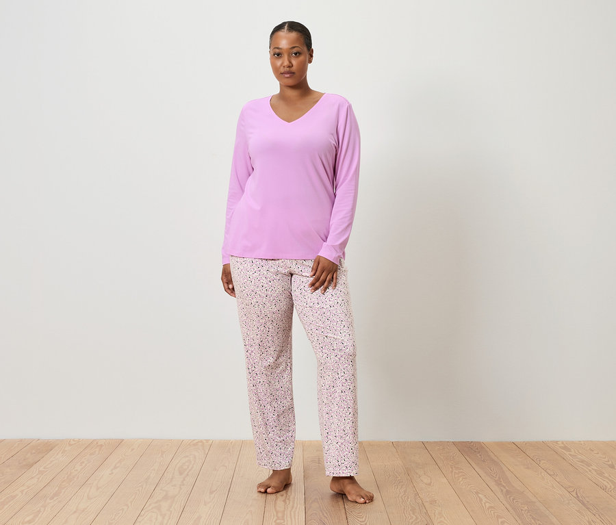 Beyaz bir duvarın önünde çıplak ayakla duran, lila pijama üstü ve çiçek desenli pembe pijama altı giyen bir kadın.