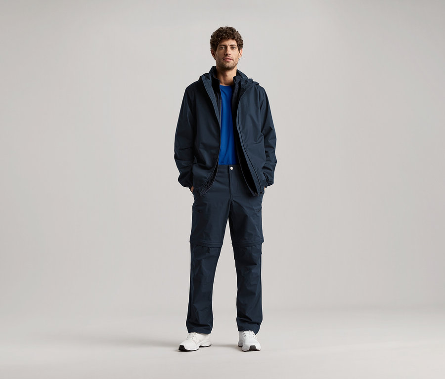 Bir adam lacivert bir ceket ve uyumlu pantolonun ceplerinde elleriyle duruyor, mavi bir tişört ve beyaz spor ayakkabılar giyiyor.