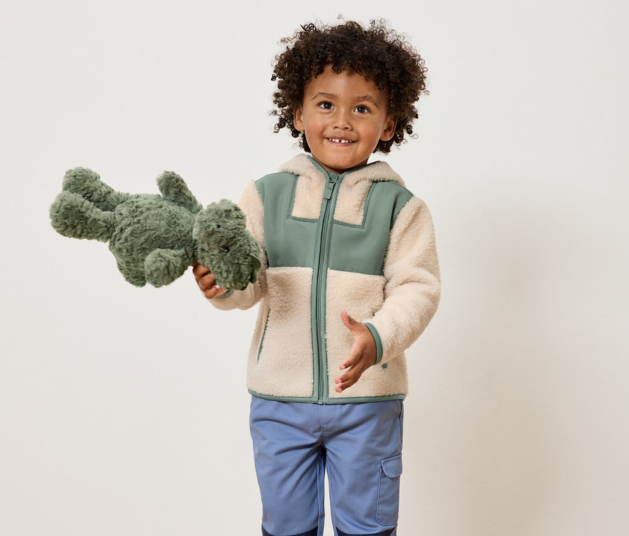Kıvırcık saçlı gülümseyen bir çocuk, yeşil bir peluş dinozor tutuyor ve yeşil detaylı açık kahverengi bir ceket ve mavi pantolon giyiyor.