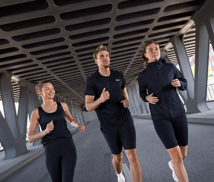 İki kadın ve bir erkek olmak üzere üç kişi bir köprünün altında koşuyor. Soldaki kadın siyah bir Spor Atlet ve siyah Spor Tayt "ActivePower" giyiyor. Ortadaki erkek siyah bir Spor Tişörtü ve siyah Spor Şortu giyiyor. Sağdaki kadın siyah Rüzgarlık "Packable" ve siyah Spor Şortu giyiyor.