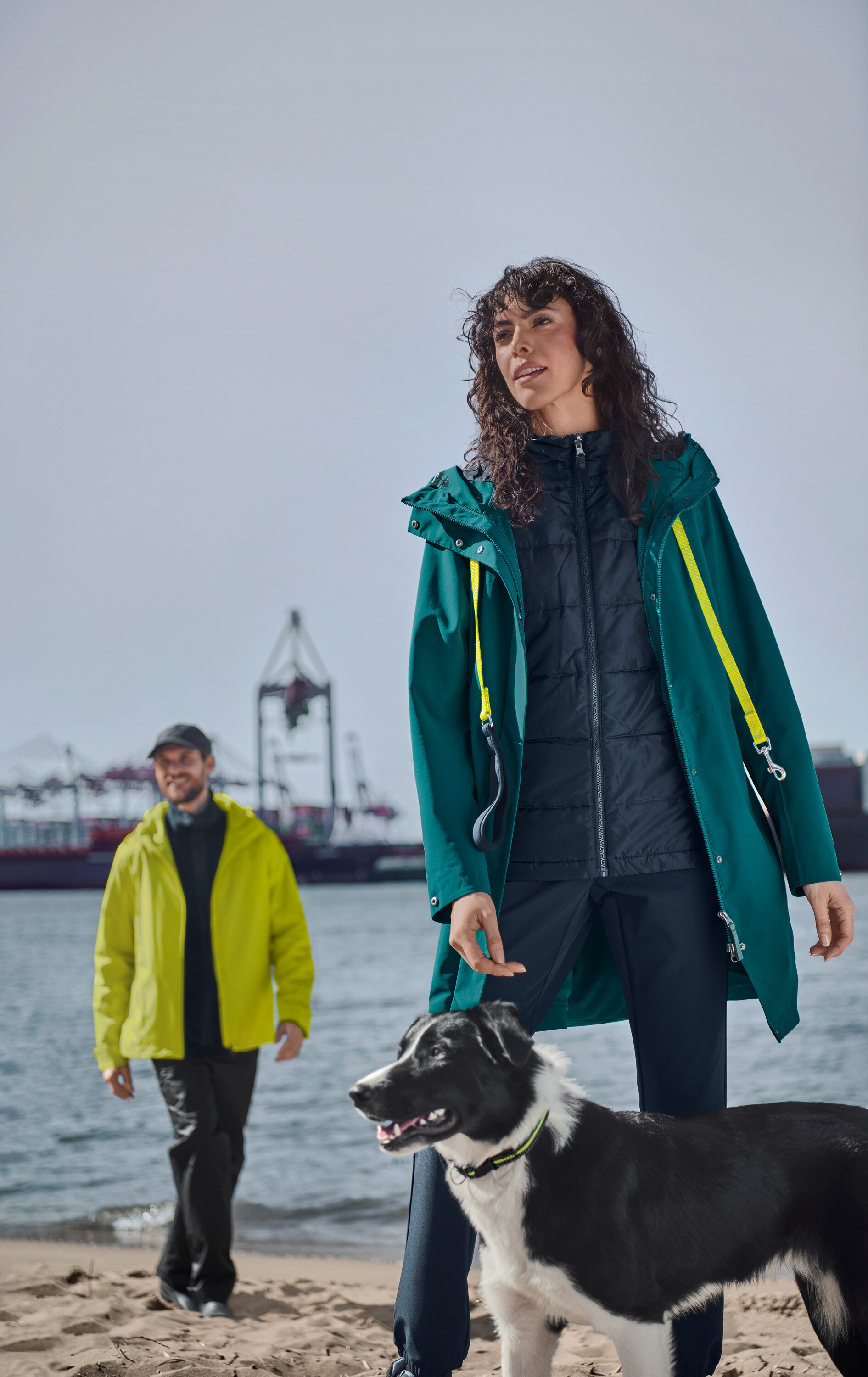 Bir kadın sahilde köpeğini gezdiriyor. Üzerinde petrol rengi 3'ü 1 arada yelekli outdoor palto ve softshell jogging pantolon var. Arka plandaki adam sarı bir yağmurluk ve kep giyiyor.