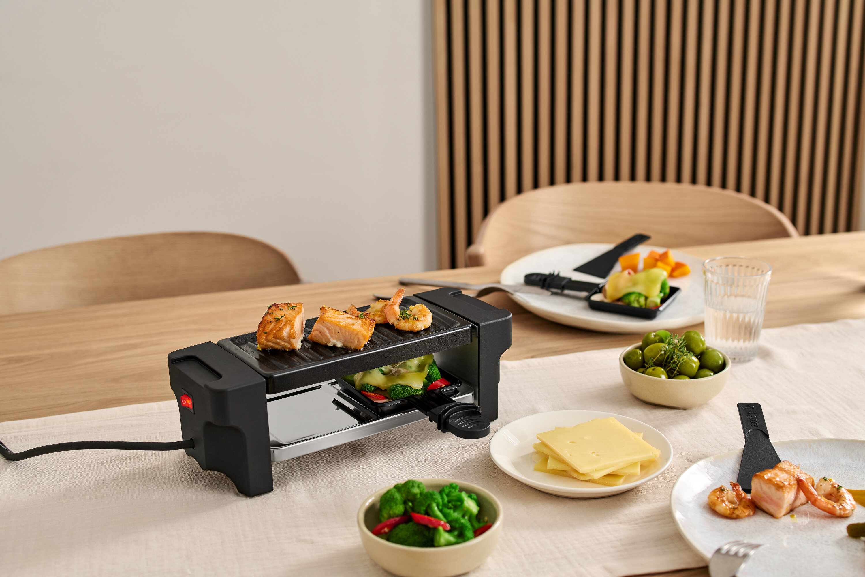 Masa üzerinde 2 Kişilik Raclette Izgara, ızgarada somon ve karidesler var. Malzemelerle dolu tabaklar ızgaranın etrafında duruyor.