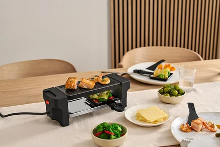 Masa üzerinde 2 Kişilik Raclette Izgara, ızgarada somon ve karidesler var. Malzemelerle dolu tabaklar ızgaranın etrafında duruyor.