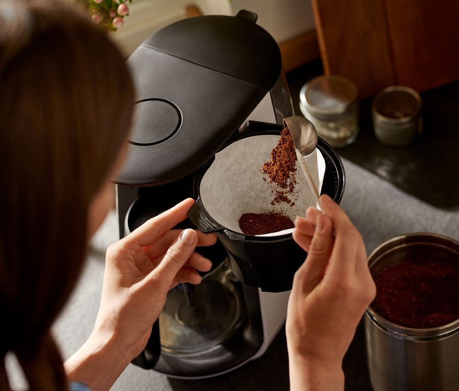 Kadın, »Let's Brew« Filtre Kahve Makinesi'nin beyaz filtresine kaşıkla kahve koyuyor.