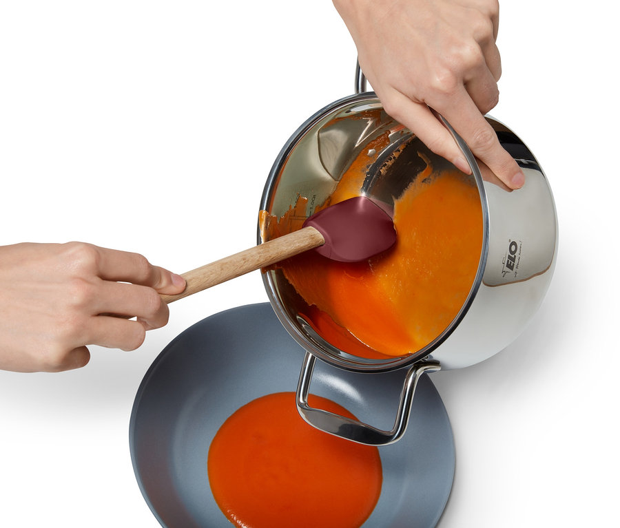 Bir kişi paslanmaz çelik tencereden gri bir kaseye turuncu bir sıvı dökerken Çok Fonksiyonlu Pişirme Kaşığı kullanıyor.