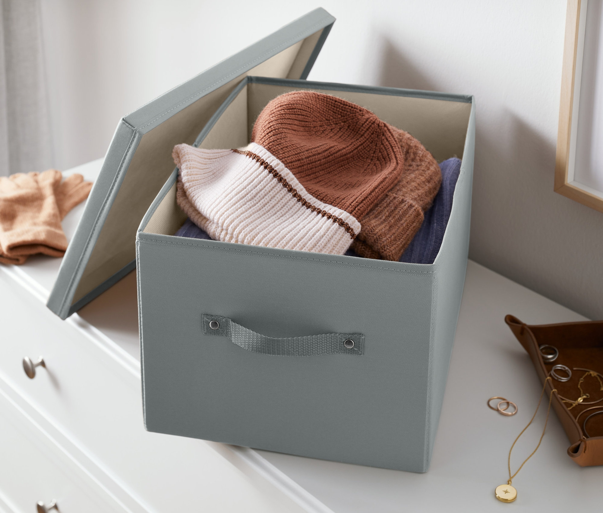 Komodinin üzerinde, içinde şapka ve kazakların bulunduğu açık gri kapaklı düzenleme sandığı duruyor.