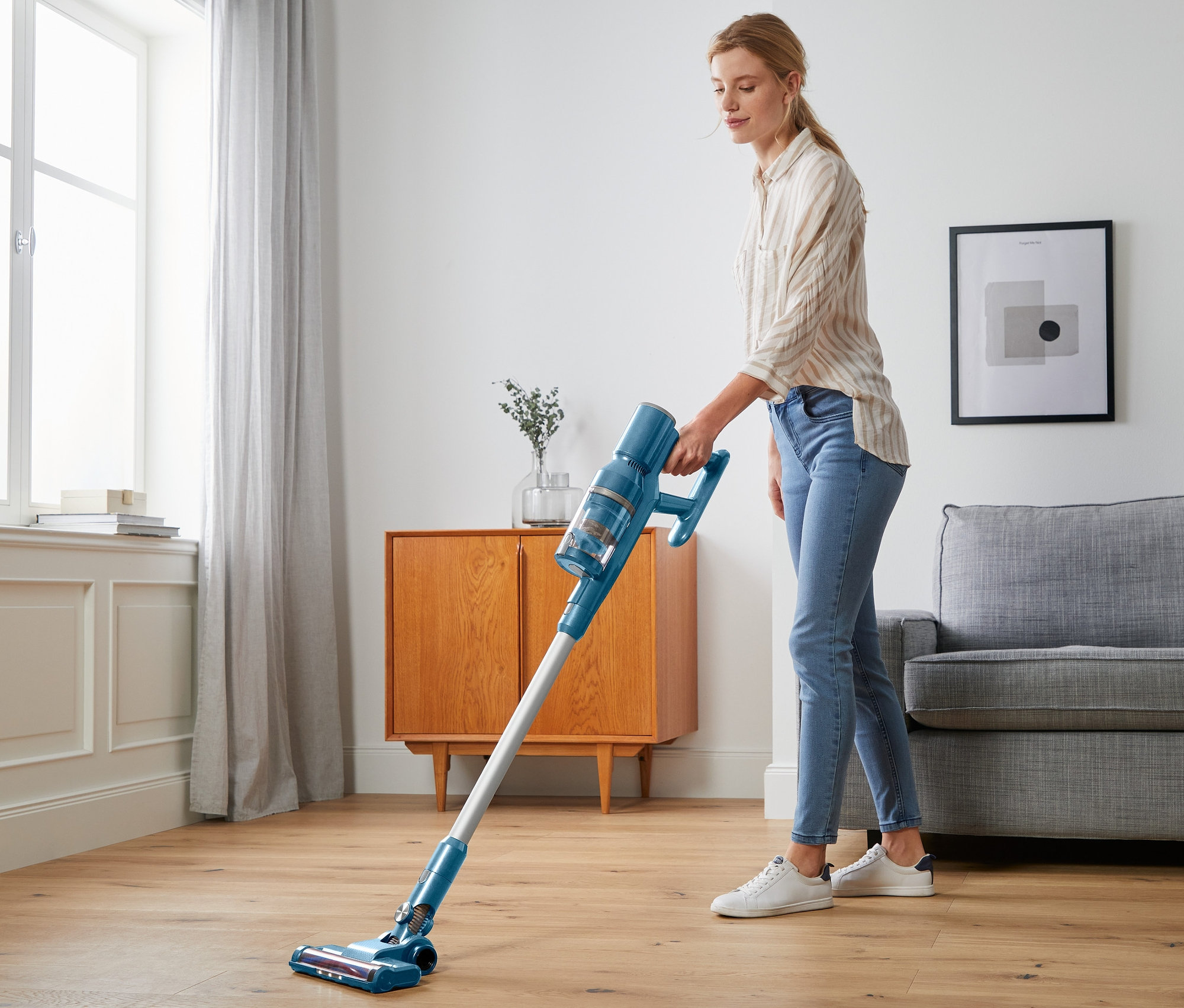 Kadın, mavi dijital kablosuz elektrikli süpürgeyle zemini süpürüyor.