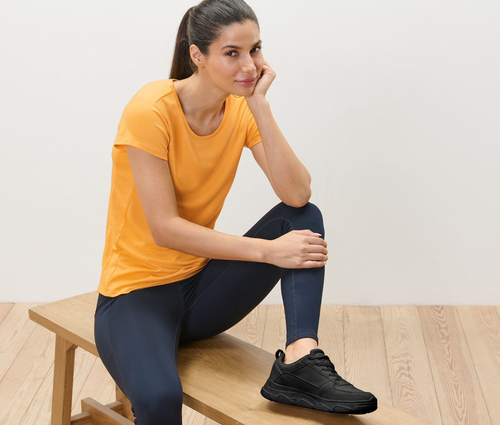 Kadın bir bankta oturuyor, turuncu bir fonksiyonel tişört giyiyor.