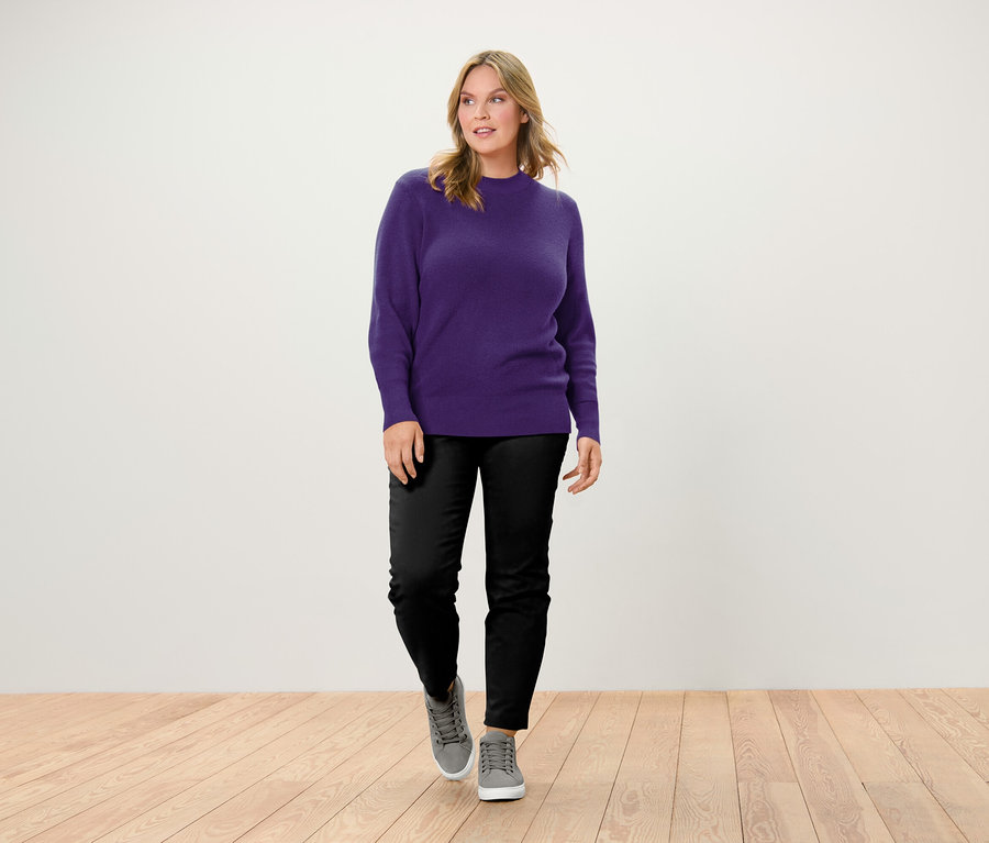 Bir kadın tahta zeminde yürüyor, üzerinde mor bir kazak, siyah pantolon ve gri spor ayakkabılar var.