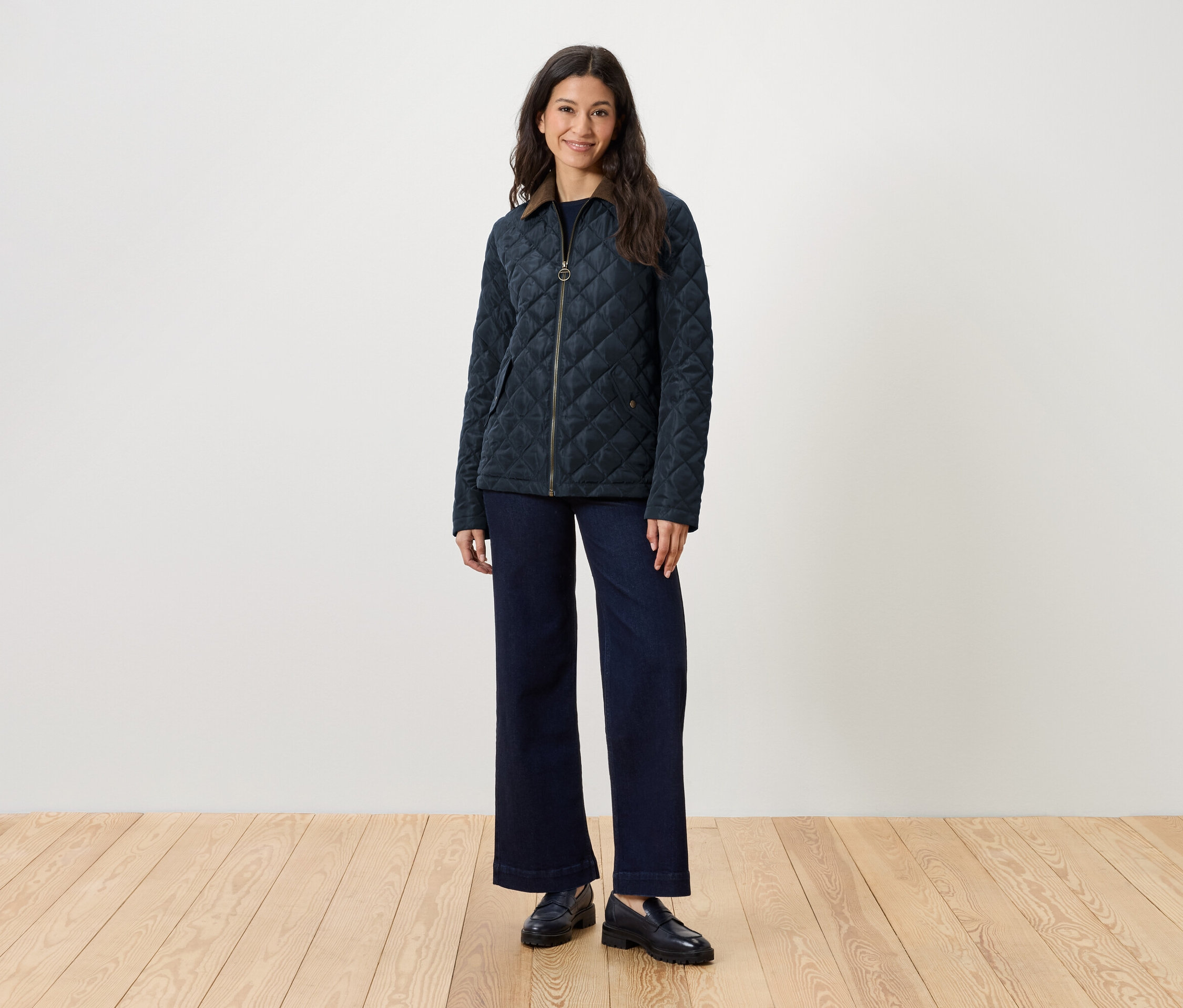 Kadın ayakta duruyor, kapitone ceket, kot pantolon ve loafer ayakkabı giyiyor.