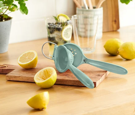 Mavi limon sıkacağı, kesilmiş limonların yanında ahşap bir kesme tahtası üzerinde duruyor. Arka planda limon ve yaban mersini bulunan bir bardak görülüyor.