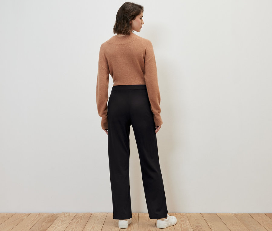 Bir kadın kameraya arkası dönük duruyor, üzerinde siyah dikişli jarse pantolon, kahverengi bir kazak ve beyaz spor ayakkabılar var.