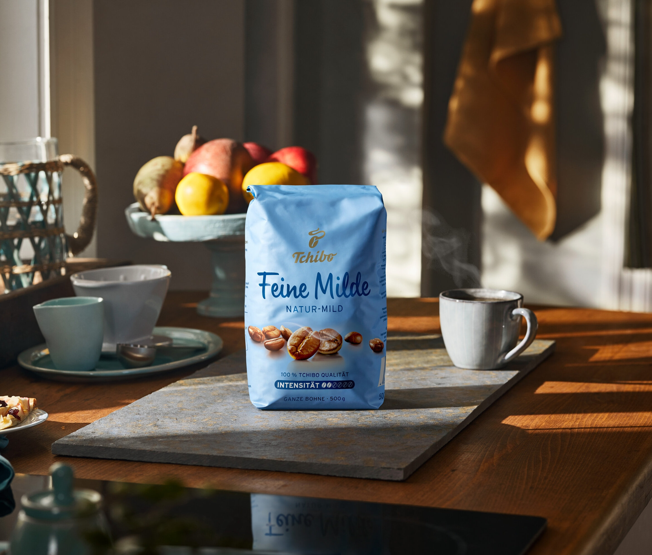 Taş bir yüzeyde bir fincan ve meyvelerin yanında 500g'lık bir paket Feine Milde kahvesi.