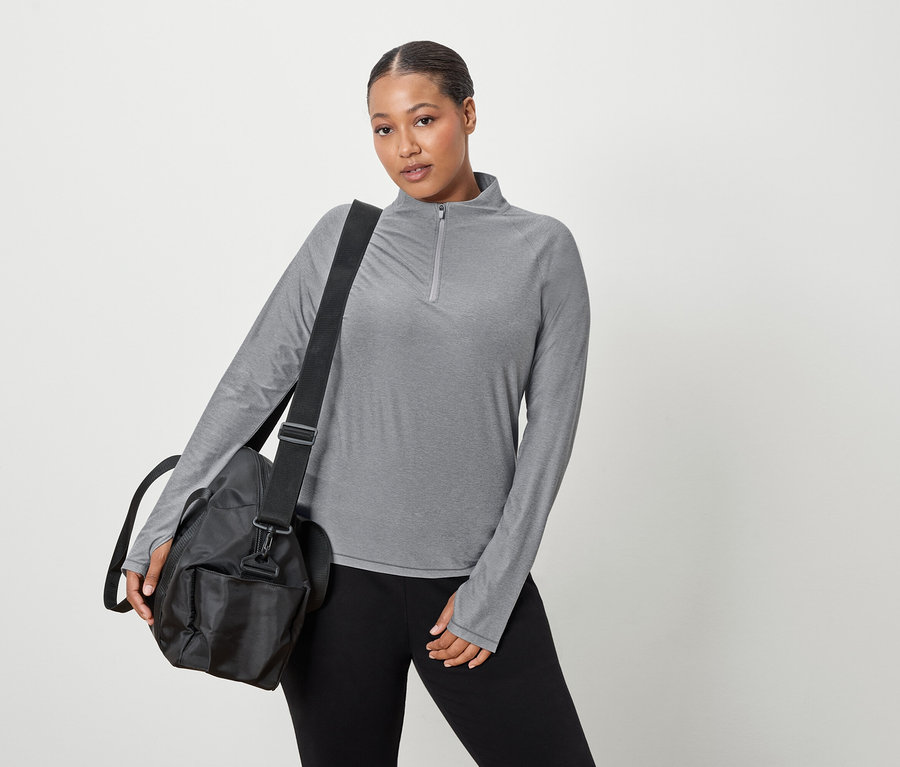 Kadın gri, uzun kollu bir üst ve omzunda siyah bir spor çantası taşıyor.