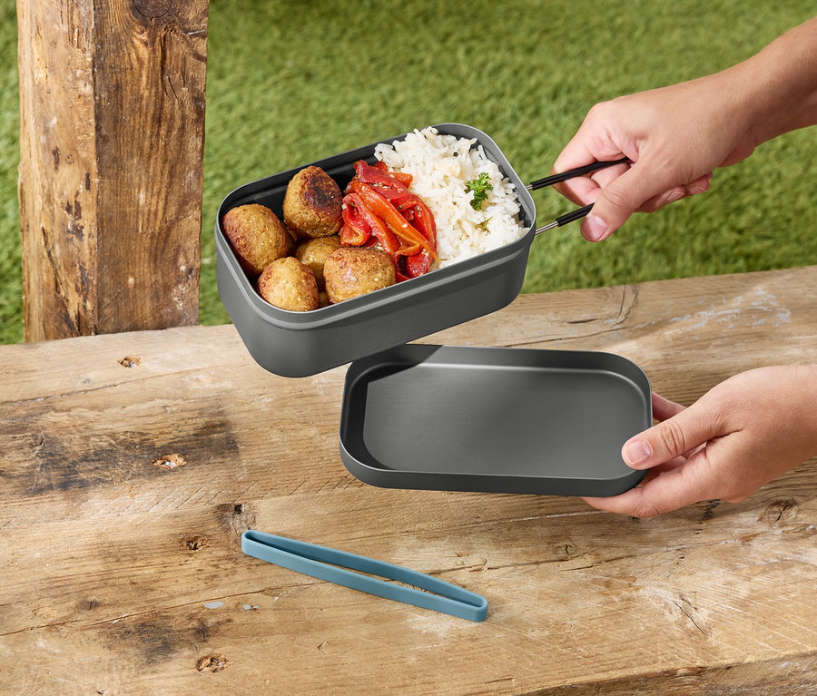 Eller ahşap bir masanın üzerinde gri kapaklı, pilavlı, köfteli ve biberli gri bir yemek kabını tutuyor.