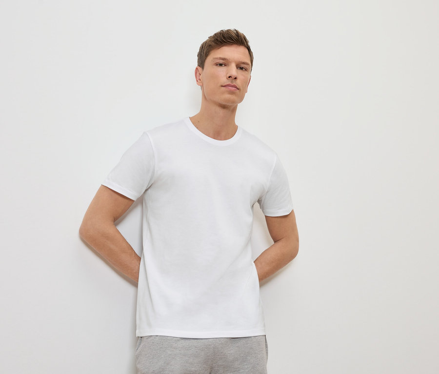 Adam beyaz bir duvara yaslanmış, beyaz bir tişört ve gri eşofman altı giyiyor.