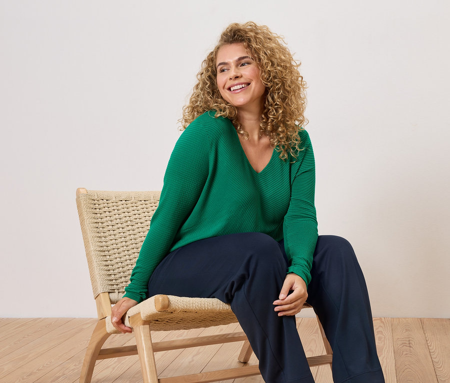 Kıvırcık saçlı bir kadın sandalyede oturuyor, yeşil bir kazak ve mavi pantolon giyiyor.