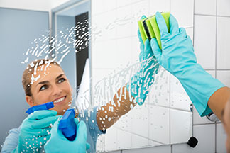 Banyo aynasının doğru temizlenmesi: Genç bir kadın aynasını mavi eldiven ve temizleme spreyi ile temizliyor.