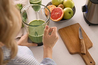 Mutfak aletleri iş başında: Bir kadın mutfakta duruyor ve içinde yeşil Smoothie olan mikseri tutuyor, yanında bir kase meyve ve üstünde bıçak olan bir kesme tahtası.