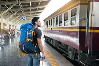Koffer für Herren oder Rucksack? Ein junger Mann hält eine Karte in der Hand, steht mit einem Rucksack an einem Bahnsteig und betrachtet den vor ihm stehenden Zug.